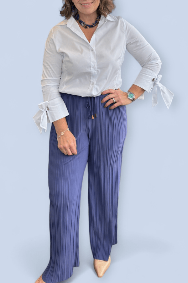 Woman wearing a white blouse with ruffled sleeves and blue pleated pants against a light blue background