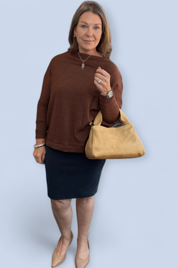 Woman wearing a brown sweater and black skirt holding a tan handbag against a light blue background