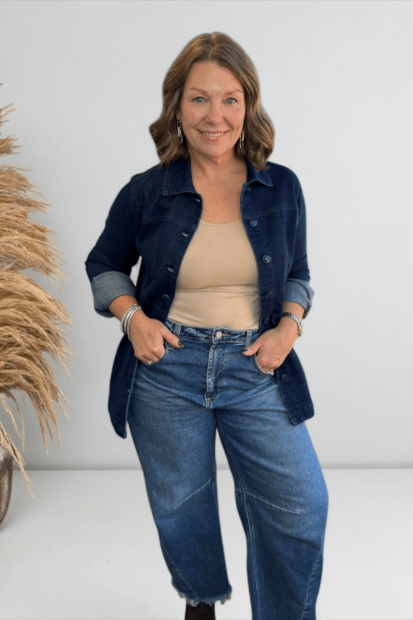 Woman wearing a navy denim jacket, beige top, and blue jeans against a white background