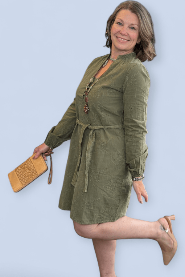 Woman wearing an olive green dress with a belt, holding a beige clutch against a light blue background