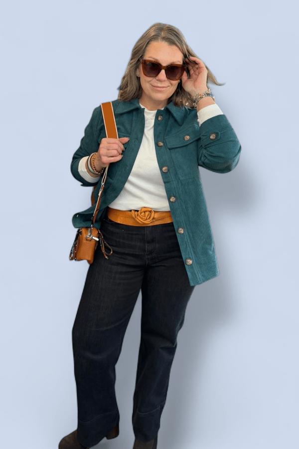 Woman wearing a teal jacket, white shirt, black pants, and brown belt against a light blue background