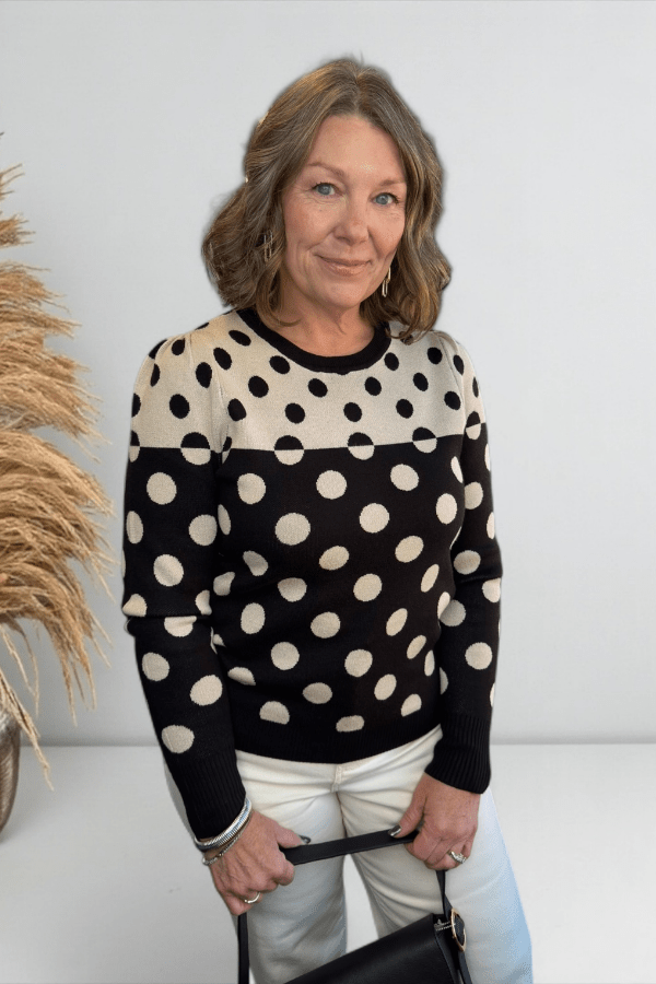 Woman wearing a black and white polka dot sweater holding a black handbag against a white background.