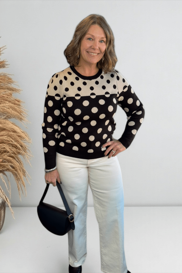 Woman wearing a black and white polka dot sweater with white pants, holding a black handbag against a white background.