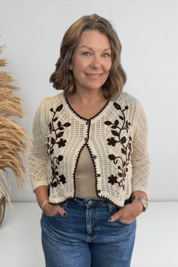 Woman wearing a cream-colored crochet cardigan with black floral patterns against a white background.