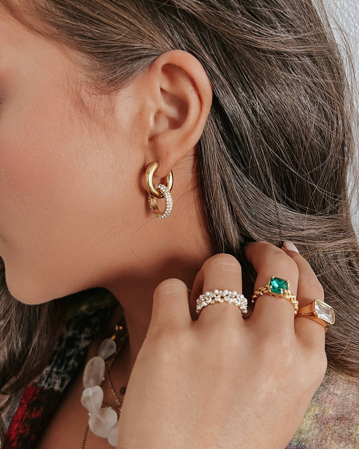 Close-up of an ear with gold hoop earrings and a hand wearing multiple rings.