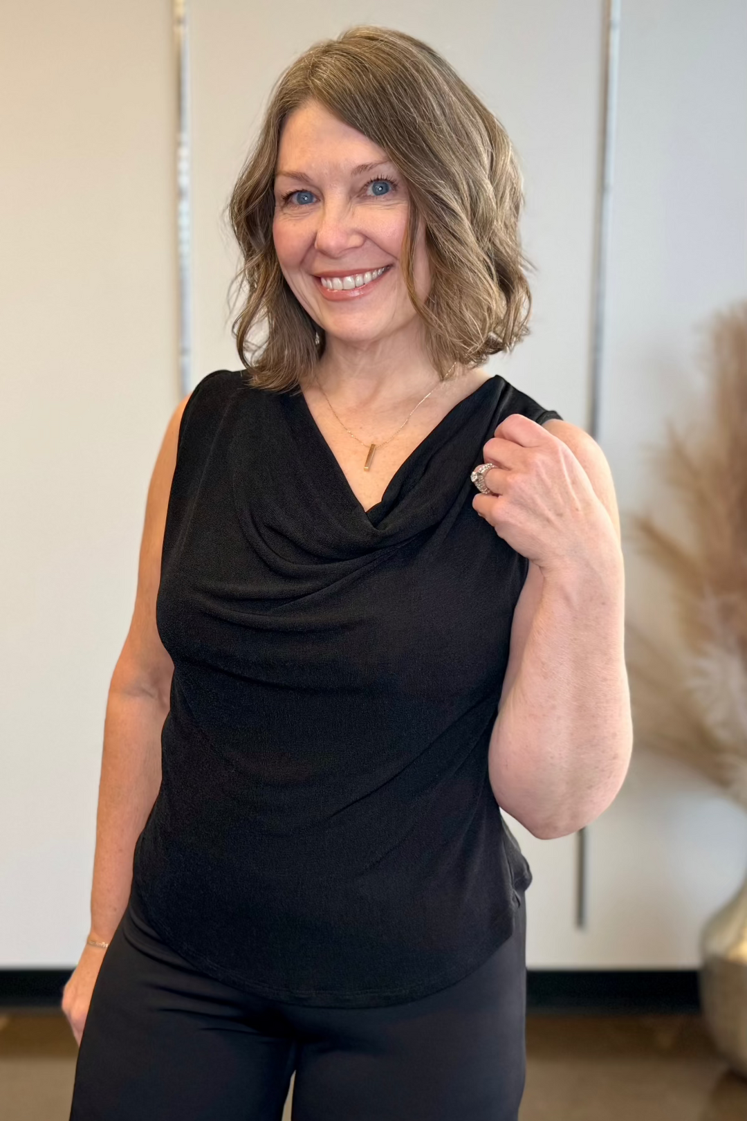 A woman smiling, wearing a black sleeveless top with a V-neckline and front cowl neckline, standing in a room with a mirror.