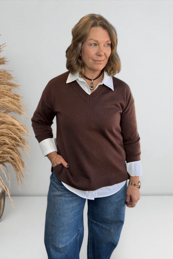 Woman wearing a brown sweater over a white shirt with blue jeans, standing against a plain background.