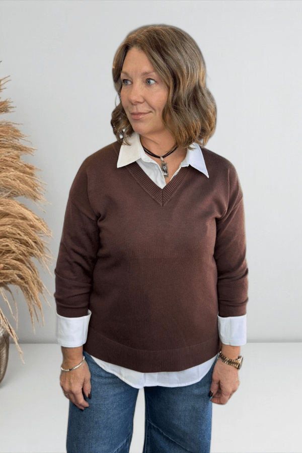 Woman wearing a brown sweater over a white shirt with blue jeans, standing against a plain background.