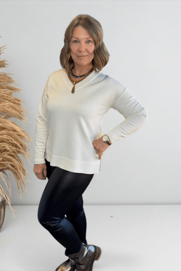 Woman wearing a white sweater and black pants standing against a plain background