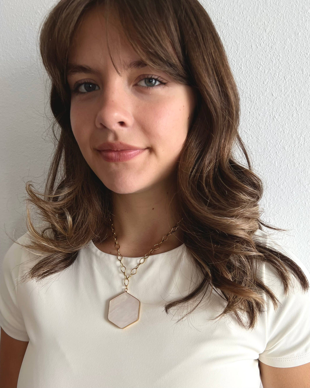 Young woman with long brown hair wearing a white shirt and a necklace with a hexagonal pendant against a plain background.