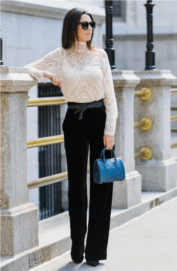 Woman in a white lace top and black pants holding a blue handbag, standing on a city street.