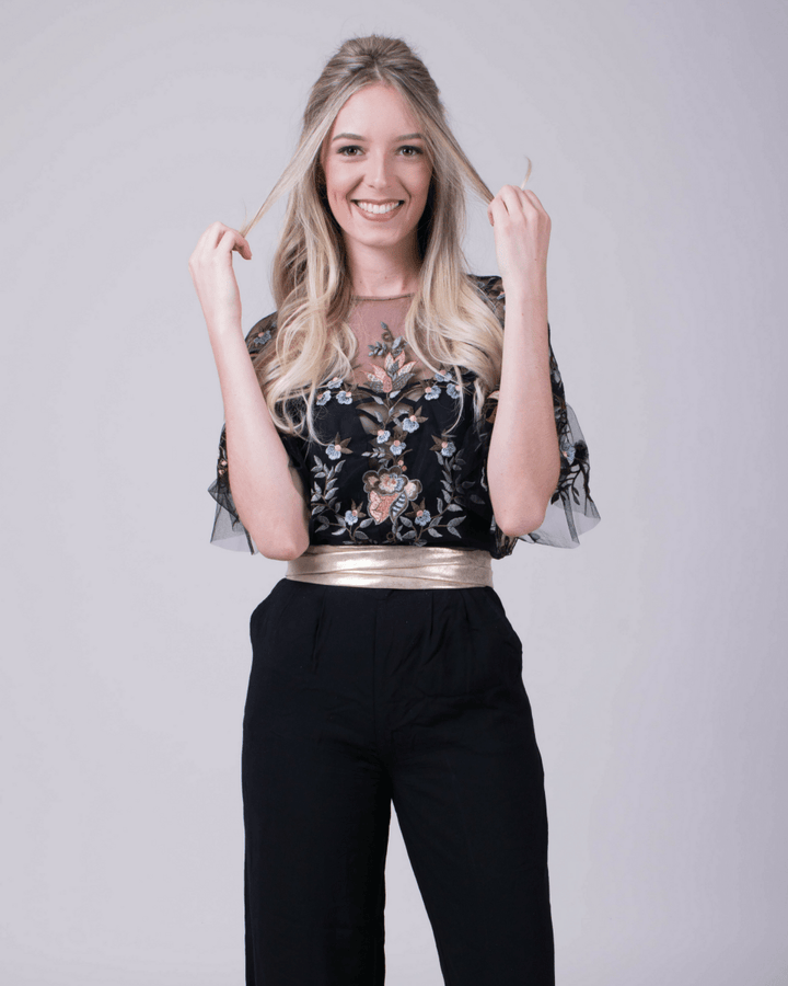 Woman wearing a black floral top with sheer sleeves and gold waistband, paired with black pants on a light gray background.