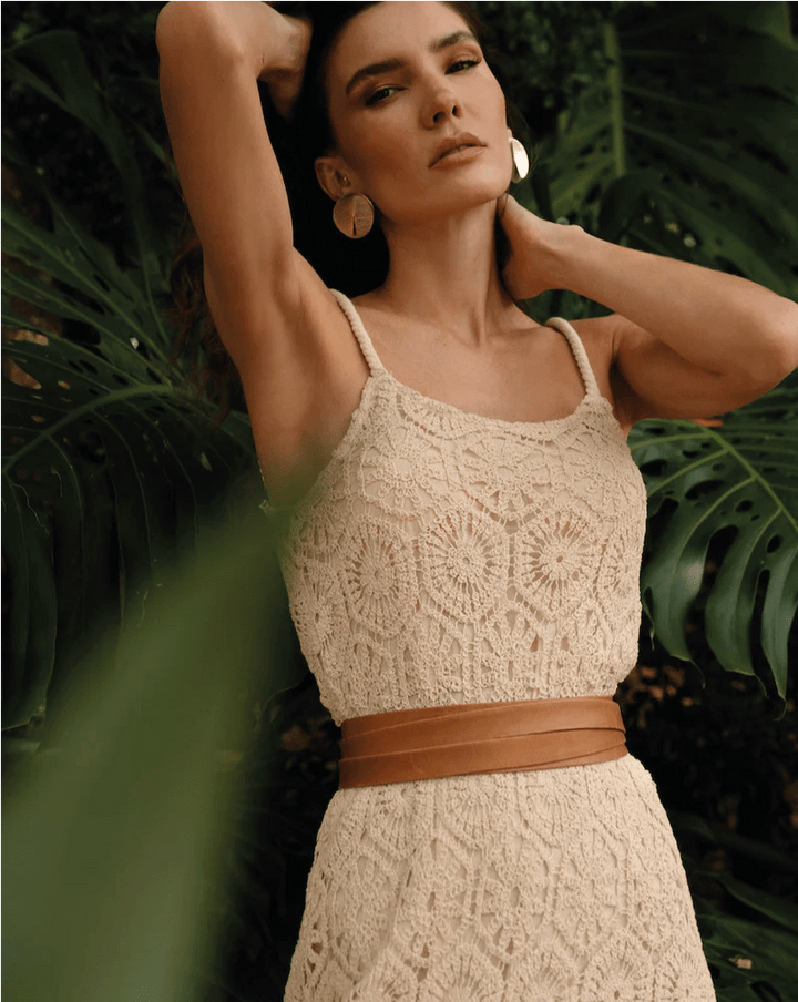 Woman wearing a beige crochet dress with a brown belt against a green leafy background