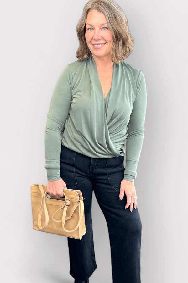 Woman wearing a green long-sleeve top and black pants, holding a beige handbag against a plain background