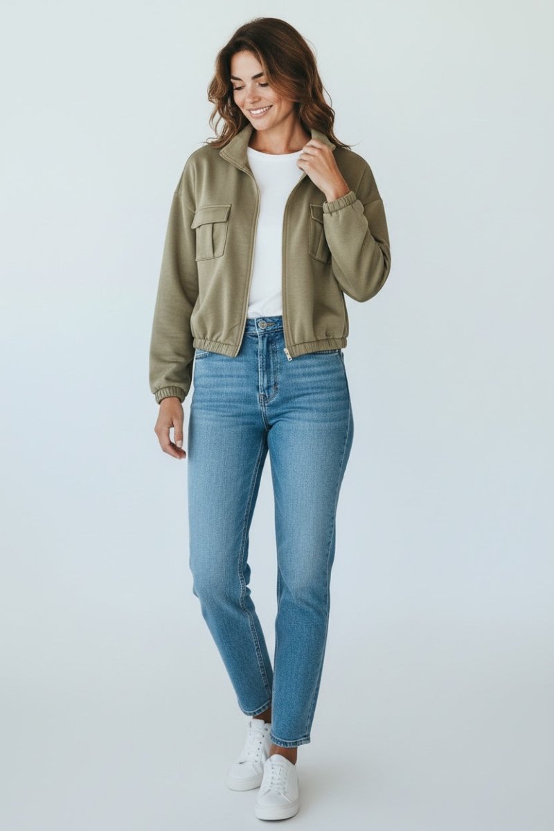 Woman wearing an olive green jacket, white shirt, blue jeans, and white sneakers on a light gray background