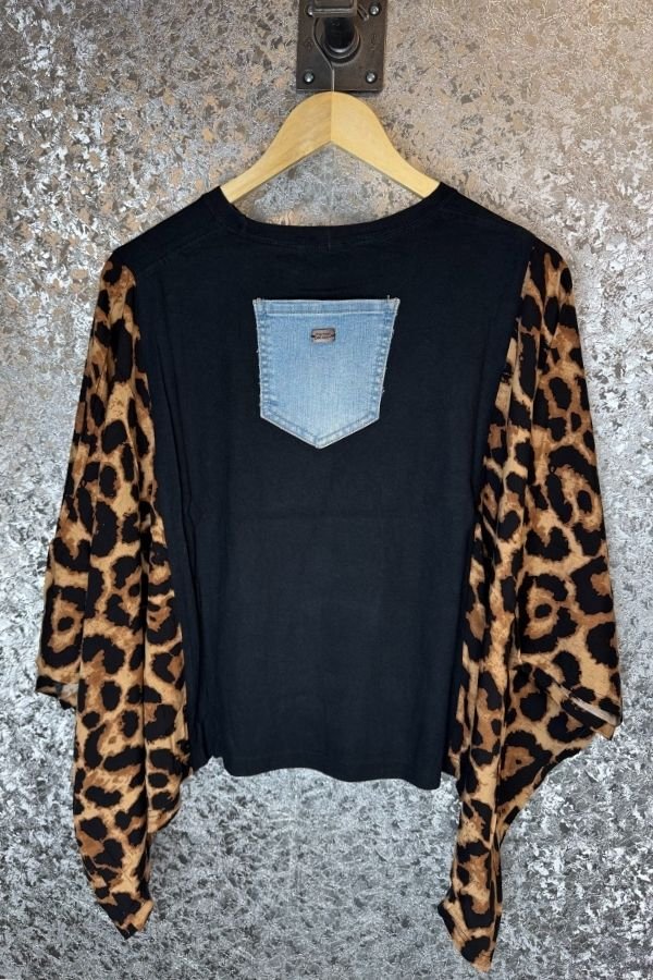 Black shirt with leopard print sleeves and denim pocket on a hanger against a textured wall.