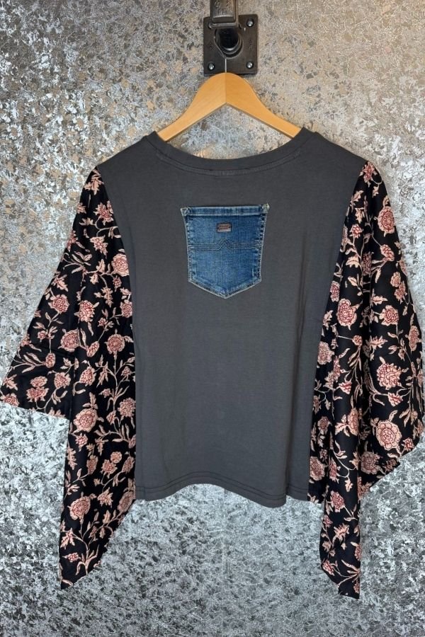Dark gray t-shirt with floral sleeves and denim pocket design, hanging on a textured wall.