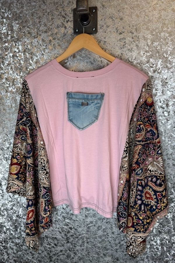 Pink shirt with denim pocket and floral sleeves on a hanger against a textured wall.
