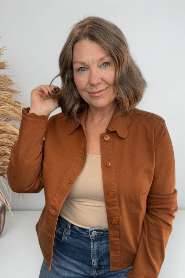 Woman wearing a brown jacket over a beige top and blue jeans, standing against a white wall with decorative elements.