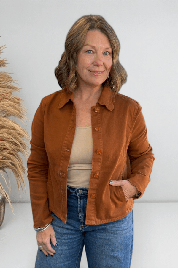 Woman wearing a brown jacket and blue jeans standing against a white wall with decorative elements.