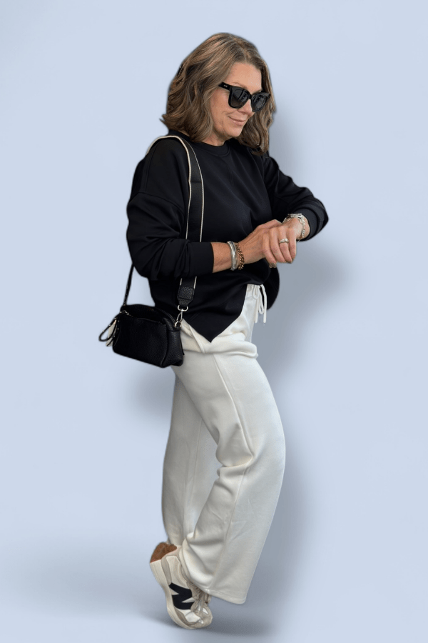 Woman wearing a black top and white pants with a black handbag against a light blue background
