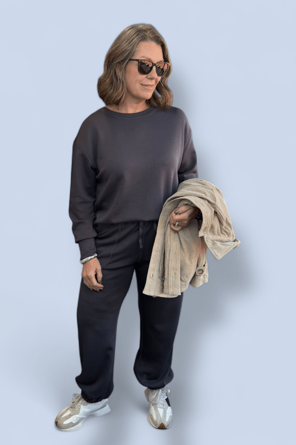 Woman wearing a dark set of joggers with a matching top and jacket held over her arm.