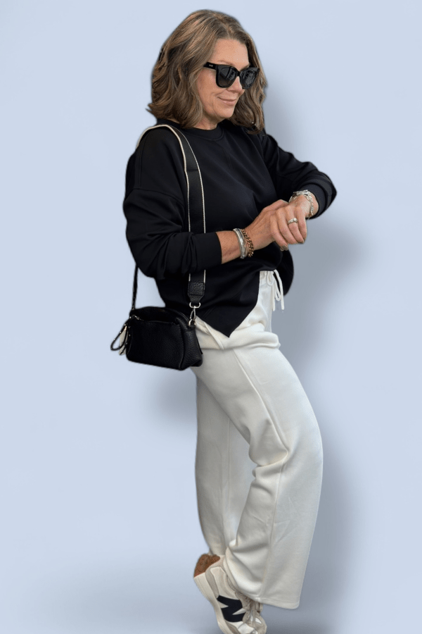 Woman wearing a black sweater and white pants with a black handbag against a light blue background
