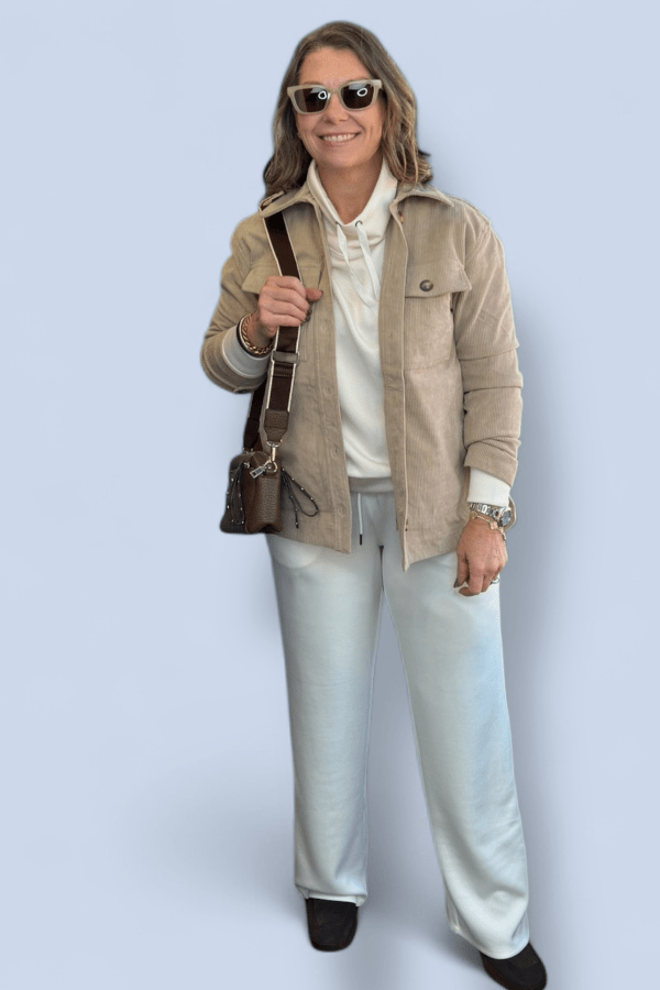 Woman wearing a beige jacket, white shirt, light blue pants, and sunglasses against a light blue background.