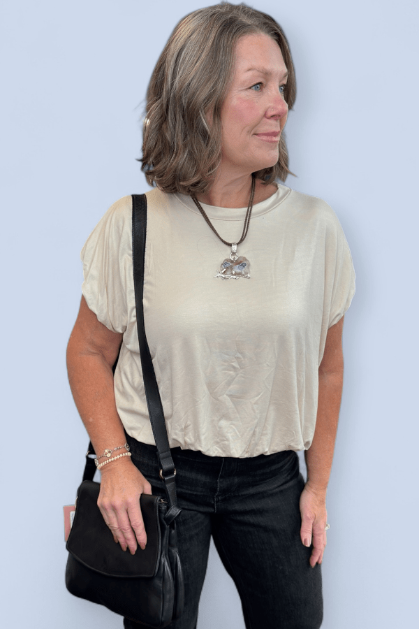 Woman wearing a beige top and black pants with a black bag against a light blue background