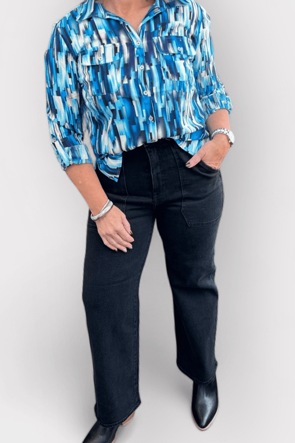 Woman wearing a patterned blue top and black jeans standing in front of white background.