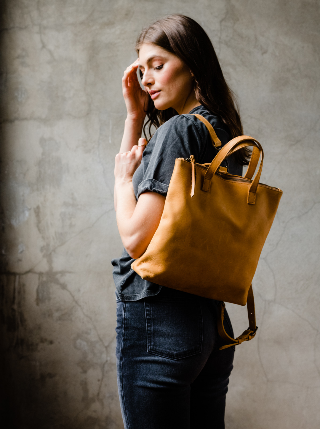 ABLE Abera Convertible Backpack in Cognac worn as a backpack with a grey linen top and dark wash jeans, side view on model