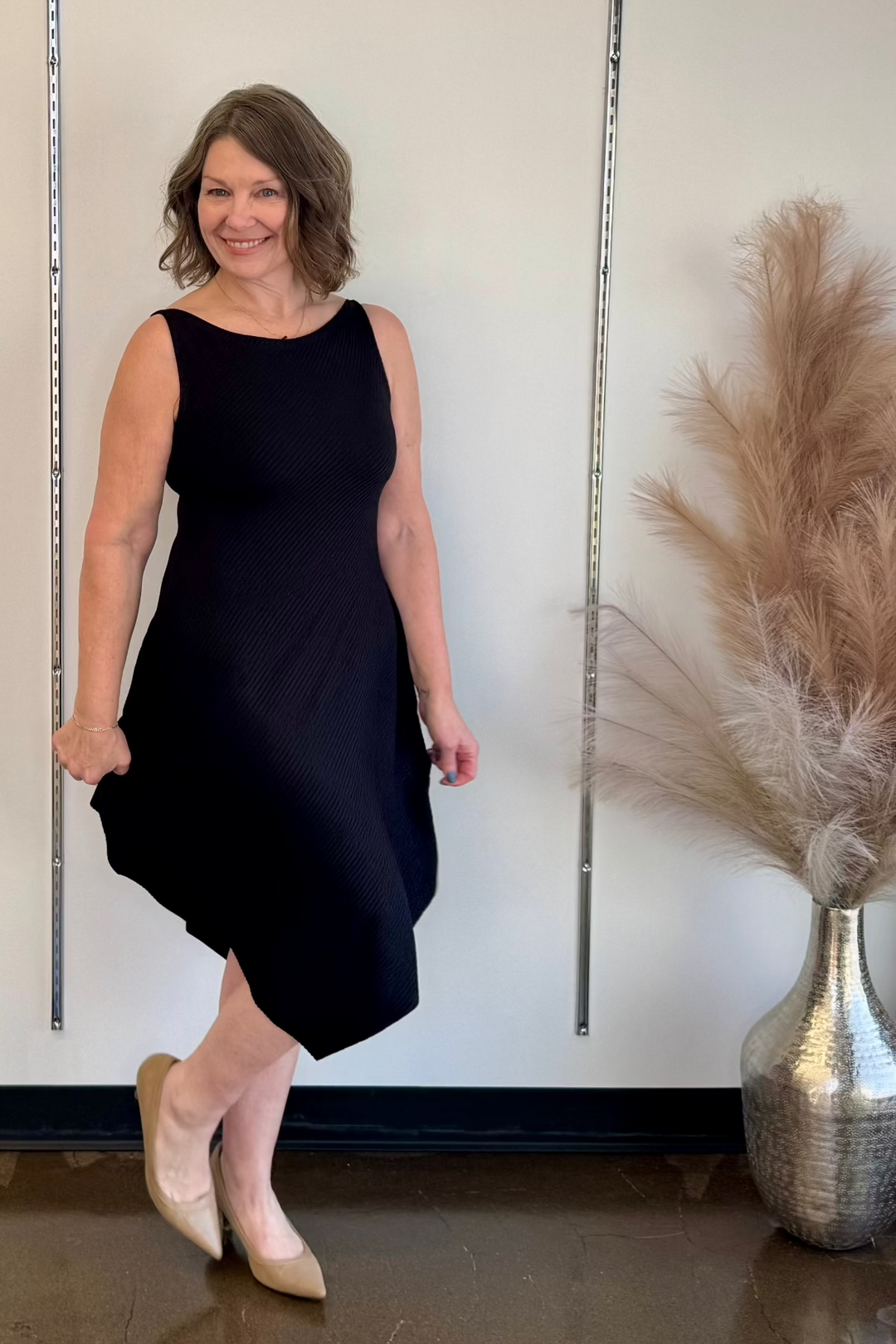A woman standing in a store wearing an elegant black knit dress with a boat neckline and sleeveless design, posing next to a metal rack with a decorative vase on it.