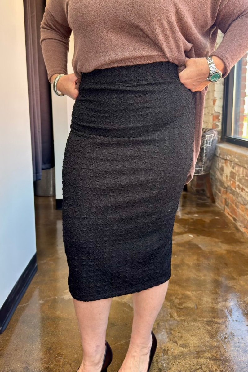 A person wearing a dark charcoal gray textured knit pencil skirt with a brown long-sleeve top, standing in an indoor setting with a polished concrete floor and brick wall.
