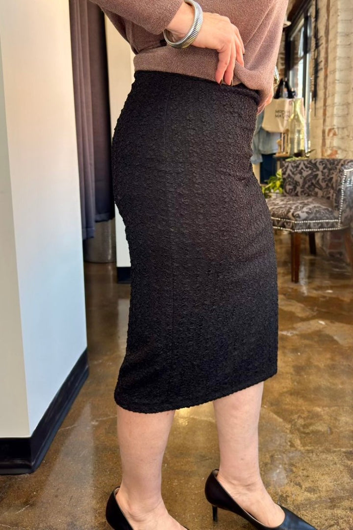 A person wearing a dark charcoal gray textured knit pencil skirt, paired with a dusty rose top and black heels, standing in an indoor boutique setting.