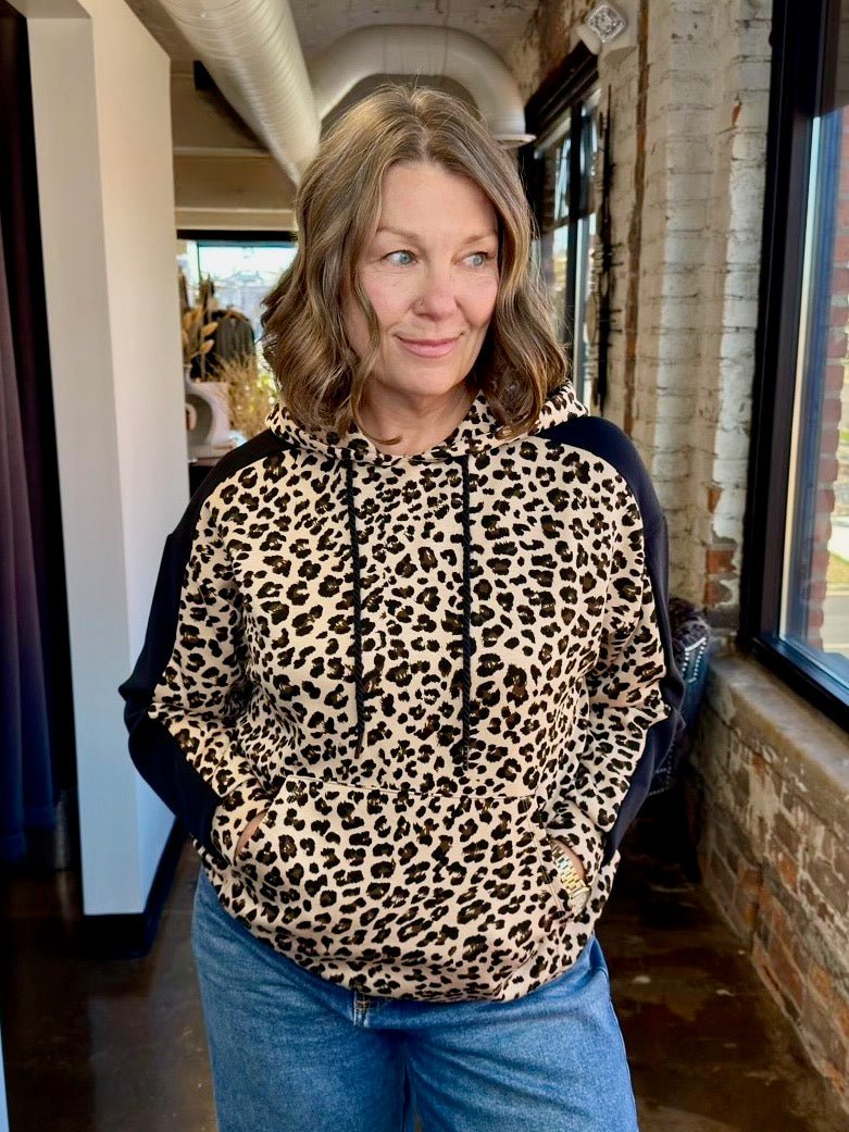 A woman wearing a leopard print hoodie with black side panels and blue jeans, standing in a brick-walled indoor space.