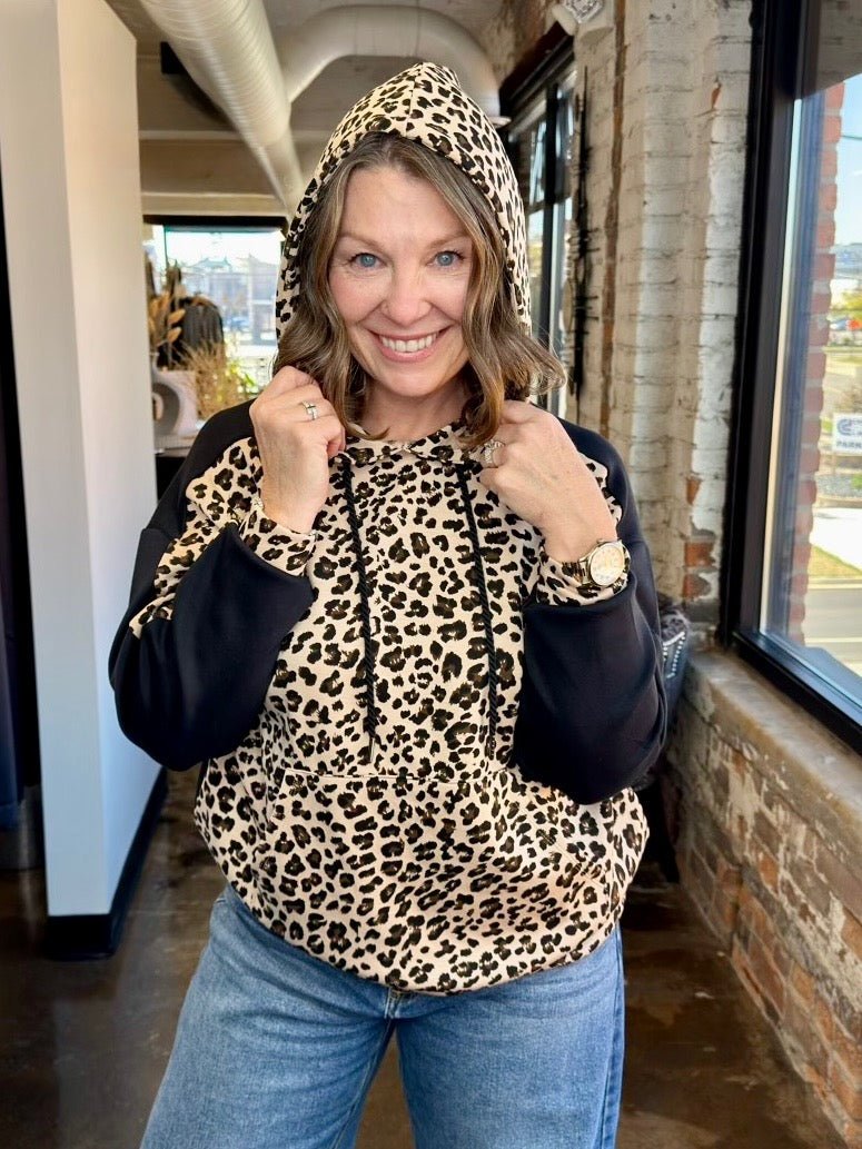 A woman wearing a leopard print and black hoodie with blue jeans, standing in a loft-style room with brick walls and windows.