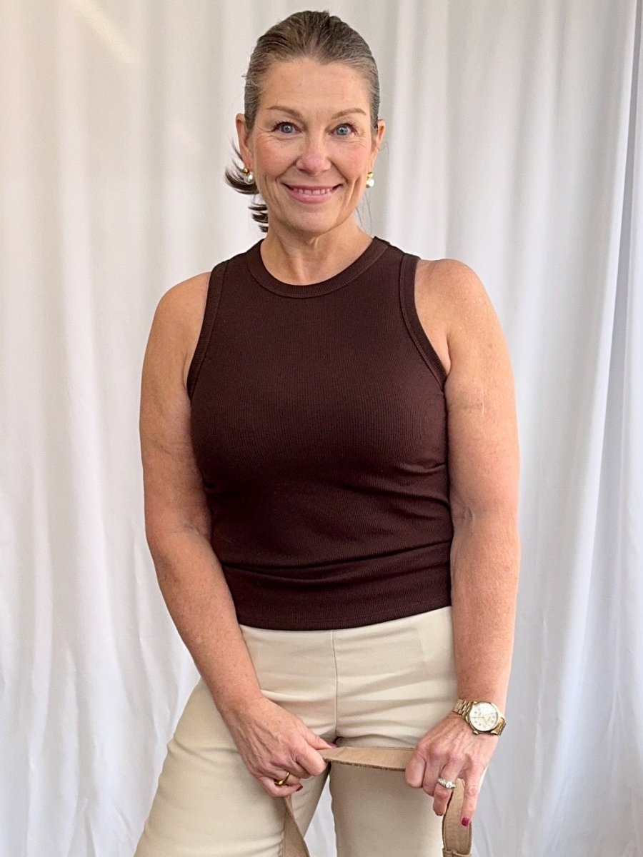 Woman wearing a brown sleeveless top and beige pants against a white curtain background