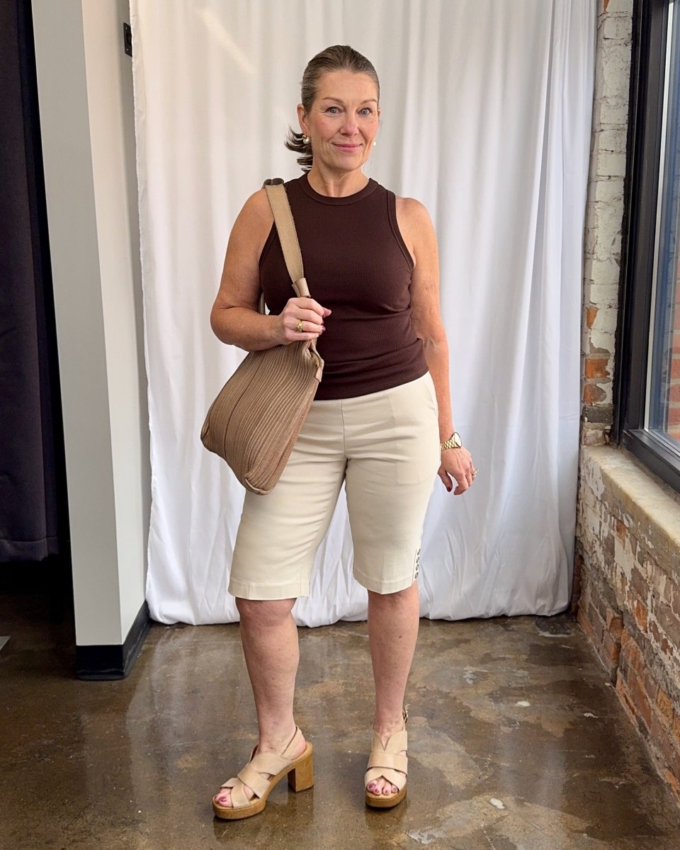 Woman wearing a brown top and beige shorts, holding a beige bag indoors.