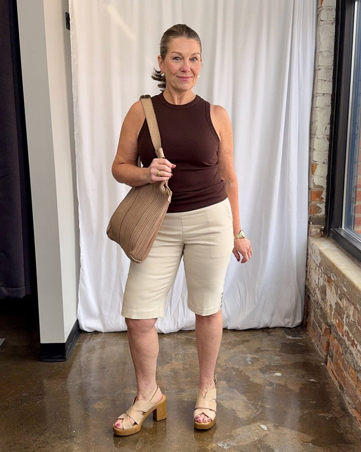 Woman wearing a brown top and beige shorts, holding a beige bag indoors.