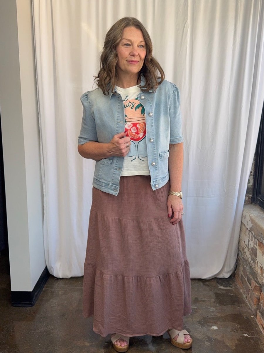 Woman wearing a denim jacket, graphic t-shirt, and tiered skirt against a white curtain background.