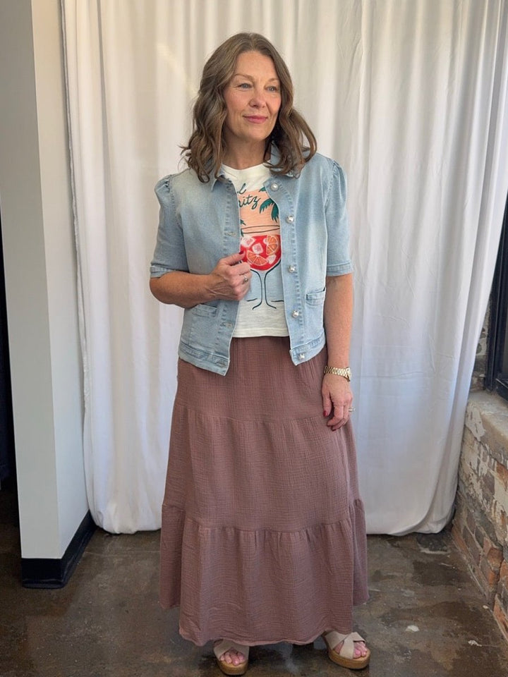Woman wearing a denim jacket, graphic t-shirt, and tiered skirt against a white curtain background.