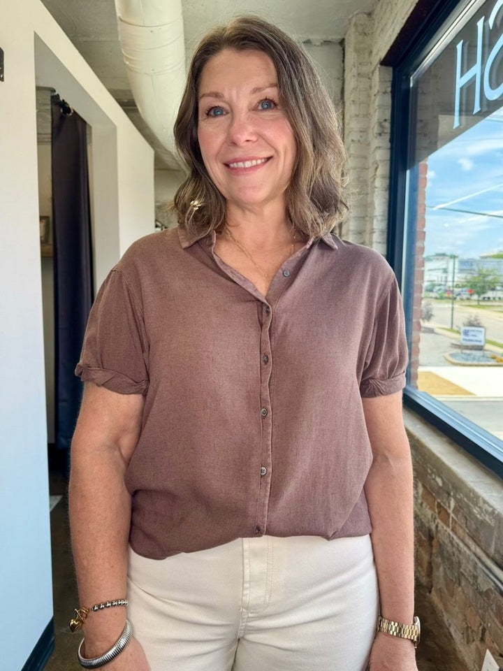 A woman wearing a short-sleeve button-up blouse in a dusty mauve-brown color, paired with off-white pants, standing in a storefront setting.