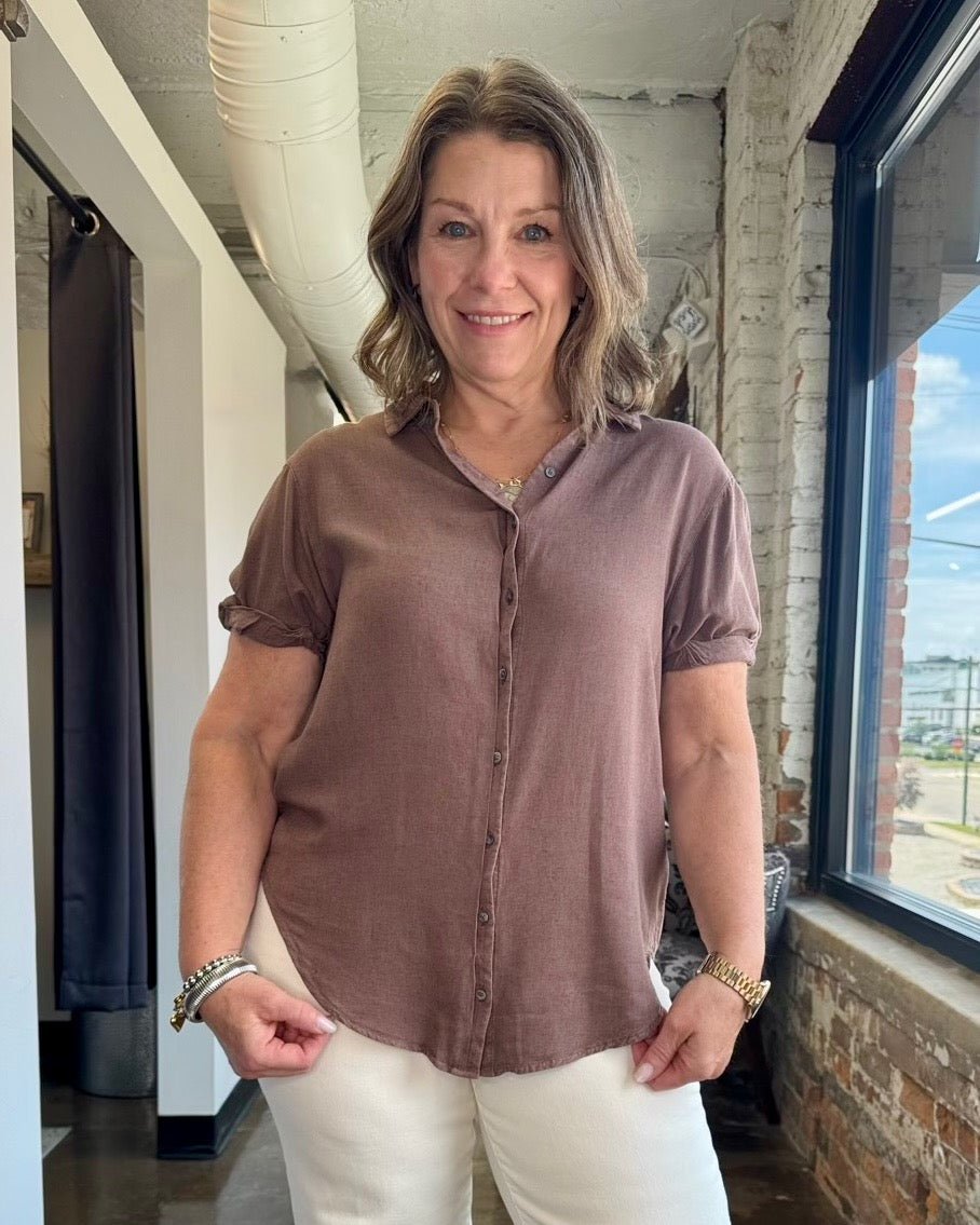 A woman wearing a short-sleeve button-front blouse in a dusty mauve-brown color, paired with white pants, standing in a loft-style room with exposed brick and a window.