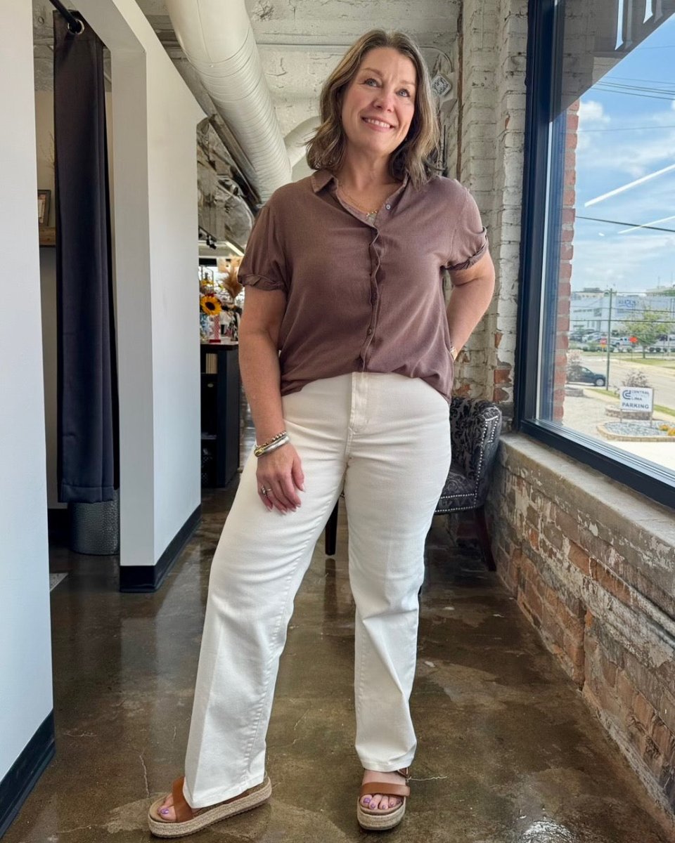 A woman modeling a short-sleeve button-up blouse in a dusty mauve-brown color paired with off-white straight-leg pants and brown platform sandals, standing in a loft-style room with a large window.