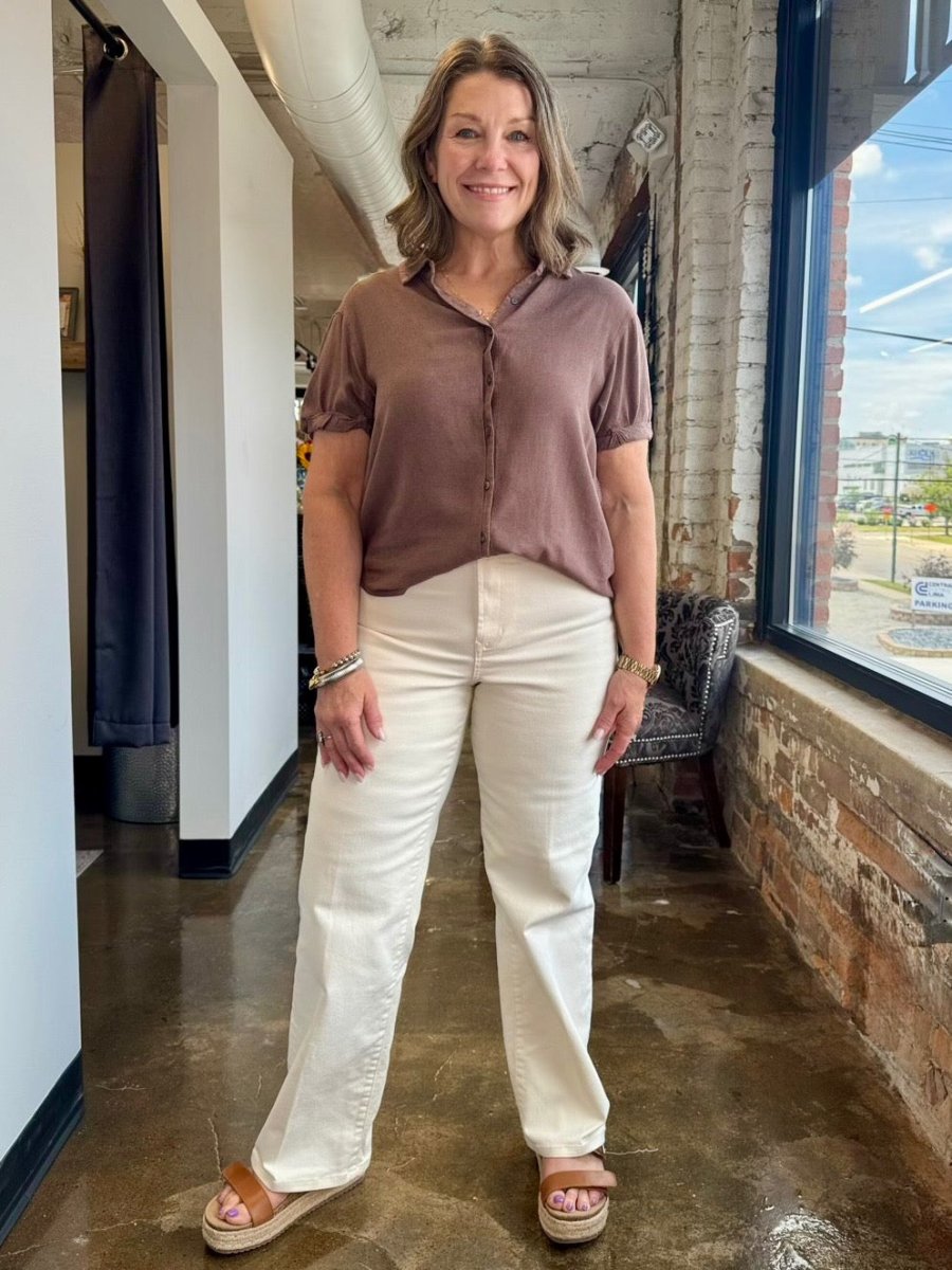 A woman modeling a short-sleeve button-up blouse in a dusty mauve-brown color paired with off-white straight-leg jeans and brown platform sandals, standing in a loft-style room with exposed brick and concrete floors.