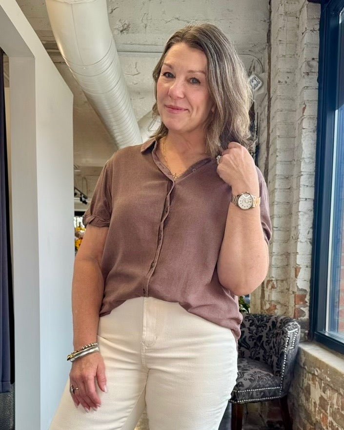 A woman wearing a short-sleeve button-up blouse in a dusty mauve-brown color and white pants, standing in a loft-style room with exposed brick and ductwork.