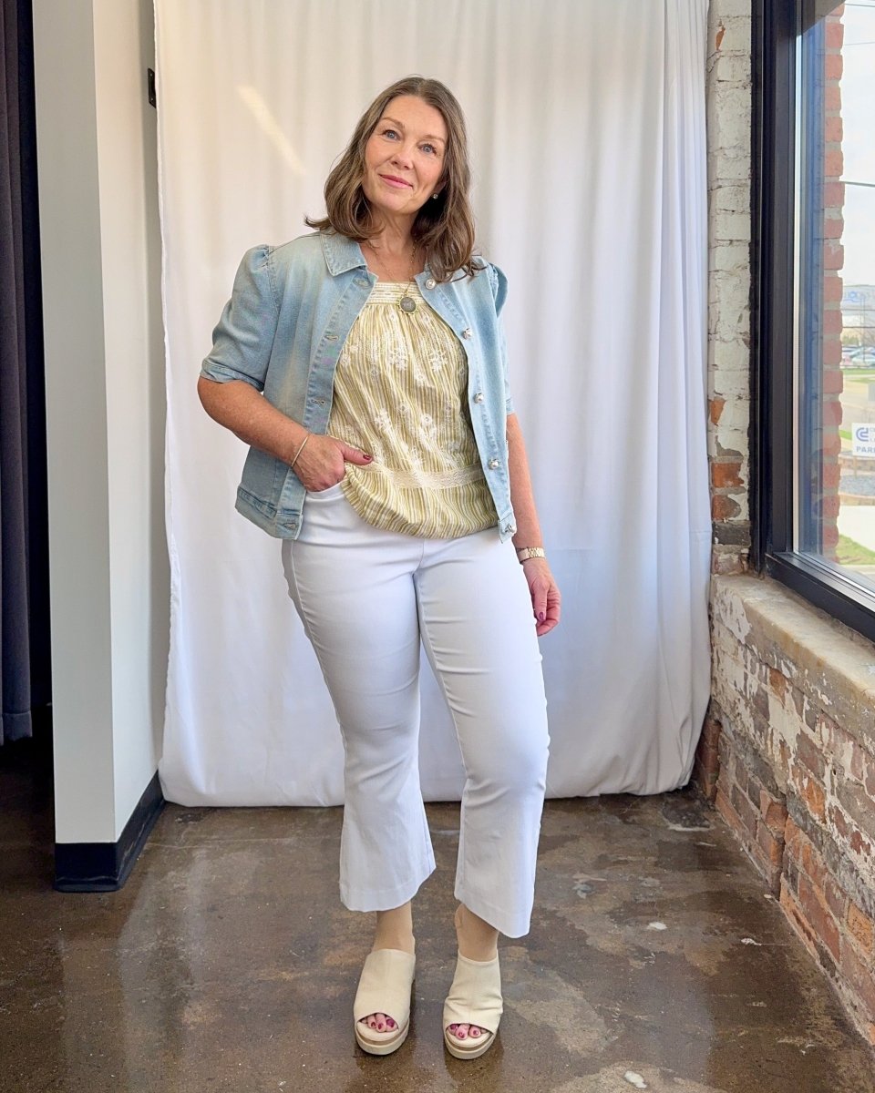 Woman wearing a light blue denim jacket over a patterned top, white pants, and beige sandals indoors.