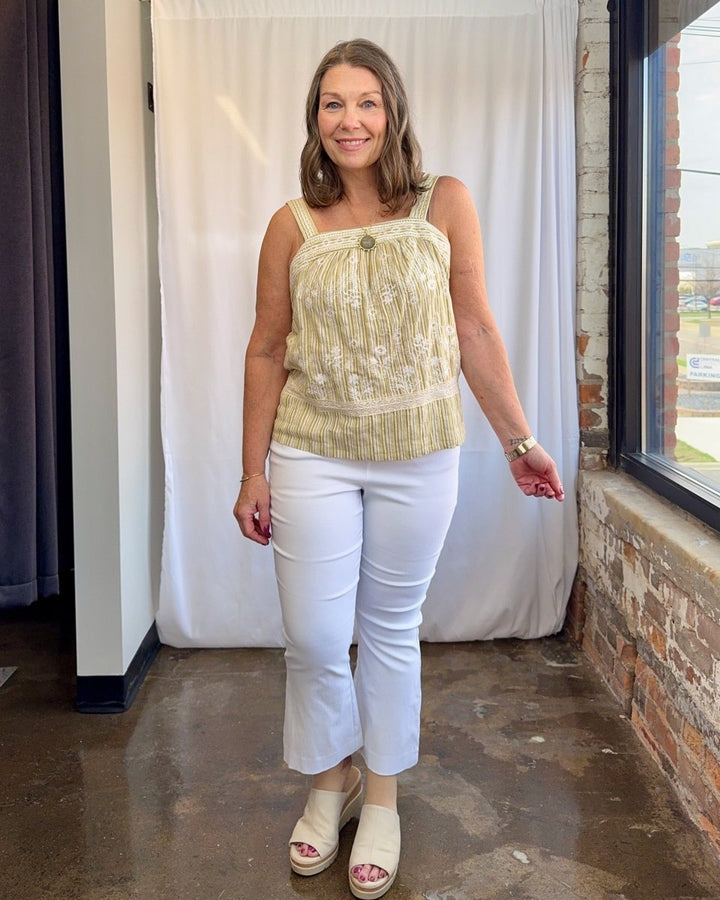 Woman wearing a yellow patterned top and white pants standing indoors.