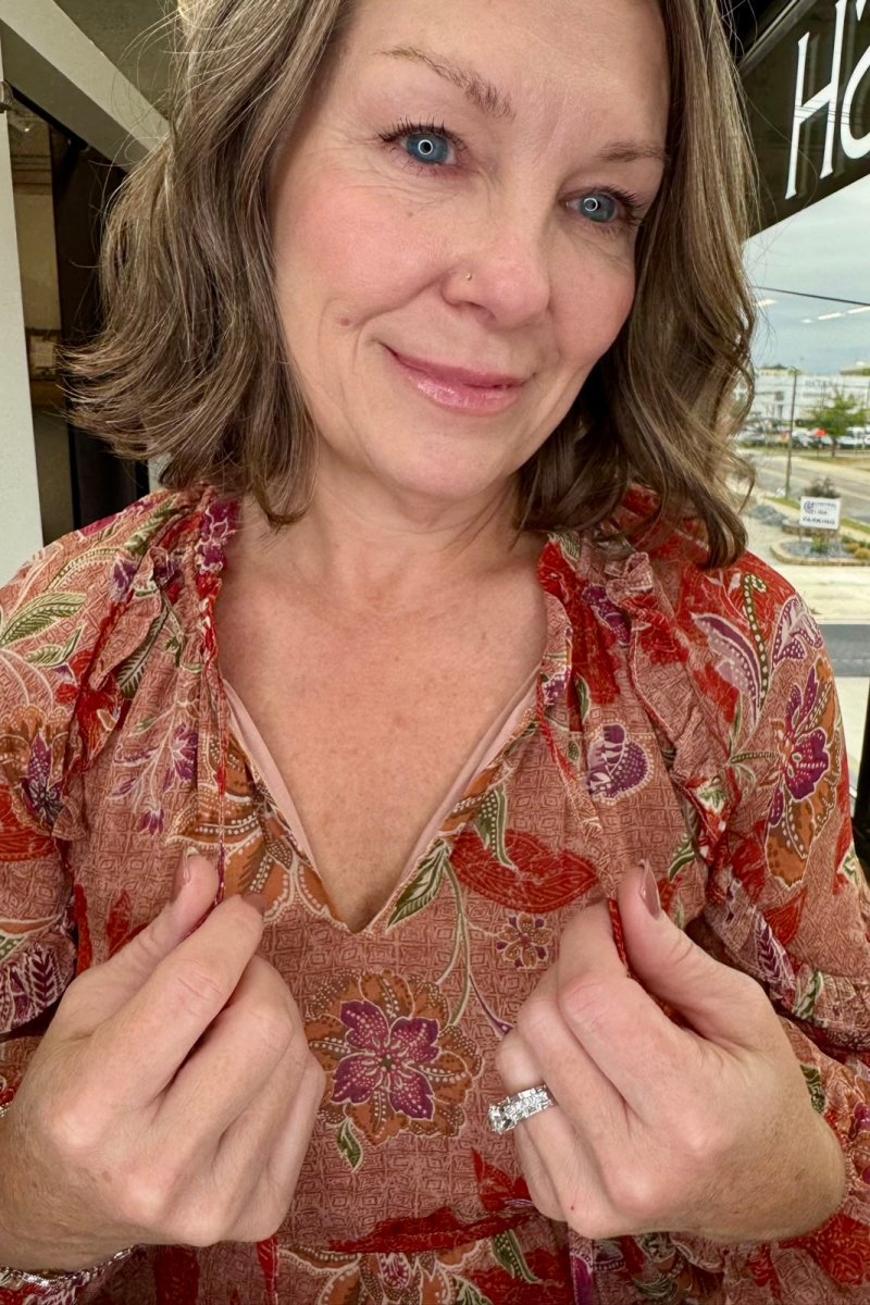 A close-up shot of a woman wearing a rust-colored floral blouse with purple and green paisley patterns, adjusting the collar of the shirt. She is wearing a silver ring on one finger, and the background shows a blurred storefront.