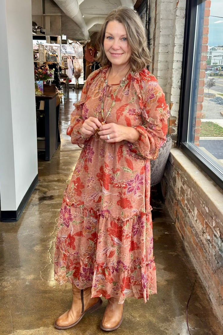A woman wearing a tiered bohemian maxi dress with a red, orange, and pink floral pattern on a sheer peach base, paired with brown leather boots, standing inside a retail store.
